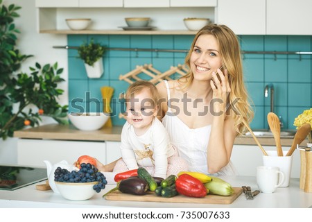 Young mother looking at camera and smiling, cooking and playing with her baby daughter in a modern kitchen. Using phone. Healthy food concept.