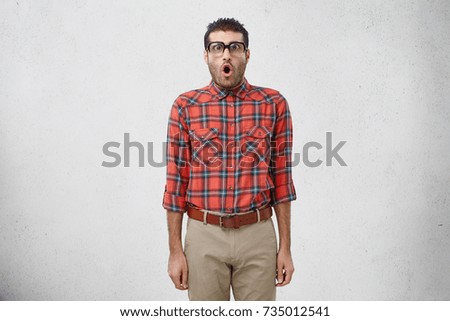 Portrait of funny shocked male geek looks at camera in terror, wears ridiculous clothes, sees something amazing in front of him. Astonished comic male nerd in square spectacles looks with bugged eyes