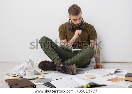Concentrated hipster guy with tattooes, sits crossed legs on floor, reads books and writes notes, being busy with studying or making project, has no spare time, prepares for session. Working concept