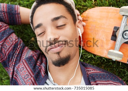 People and enjoyment concept. Close up of bearded stylish male closes eyes with pleasure, listens to favourite song in white earphones, rests on green lawn near skateboard, enjoys good atmosphere