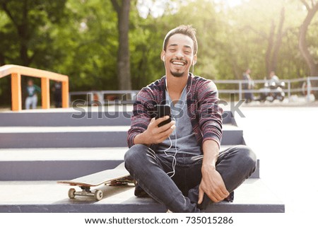 Joyful restful mixed race teenager sits on ground outdoors, uses mobile phone as listens melodies with earphones. Young talented skater rests after long training, enjoys music and sunny weather