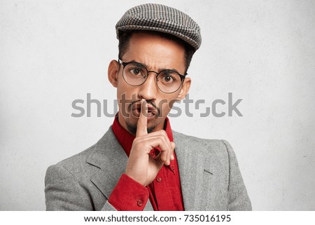 Funny male with dark healthy skin wears round spectacles, keeps finger on lips, makes silence sign, asks not to tell his secret. Bearded male wonk wants quiet atmopshere for studying or reading book