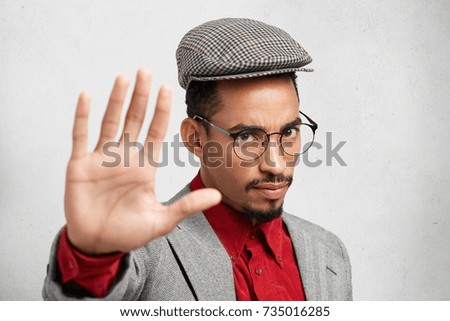 Serious dark skinned male makes stop gesture with palm, says no, expresses denial or restriction. Attractive bearded man in cap and formal jacket prevents himself from something bad by stop sign