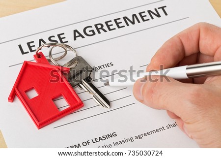 Man holding pen over house door key with red keychain pendant and lease agreement form on wooden desk - house or apartment rental concept Royalty-Free Stock Photo #735030724
