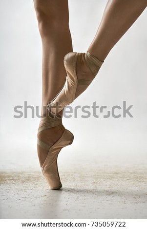 Elegant ballerina is posing in the studio on the light background. She stands on the right toe and holds left leg in the air. Girl wears beige pointe shoes. Closeup. Vertical.
