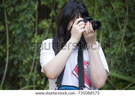 Young girl photographer with a camera in her hands taking a photo in the park in summer day in a T-shirt with a picture of a red sneaker