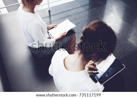 Close up of businesswoman is searching information in web via portable touch pad, while her partner is sitting near with paper documents.Young female is using digital tablet before meeting with client