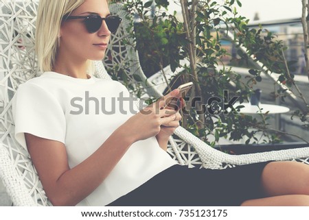 Young female student is enjoying rest and good day, while sitting with smart phone in cozy cafe terrace. Beautiful woman tourist is using cell telephone and relaxing after walking in a strange city