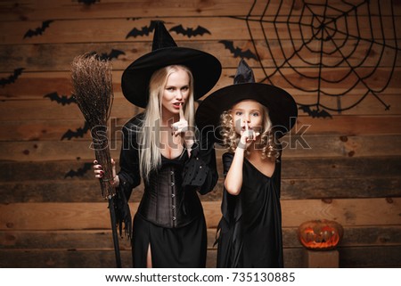 Halloween Concept - cheerful mother and her daughter in witch costumes celebrating Halloween doing silence gesture posing with curved pumpkins over bats and spider web on Wooden studio background.
