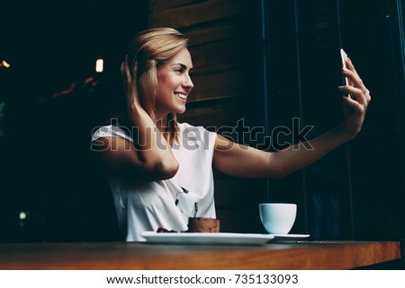Young pretty woman with beautiful smile posing while photographing herself on mobile phone camera, happy hipster girl making photo with cell telephone for a chat with friends while sitting in cafe