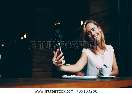 Charming happy hipster girl photographing herself on cell telephone camera while relaxing in modern coffee shop, pretty woman with cute smile making self photo on her smart phone during rest in cafe
