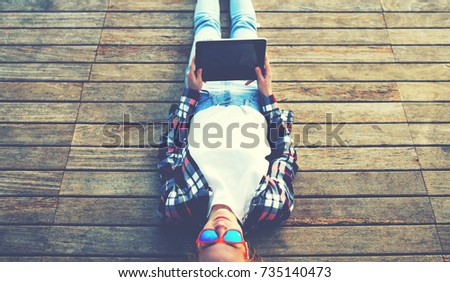 Young teen woman relaxing upon a wooden jetty while taking self portrait holding up tablet, female tourist making remembering portrait during her holidays, surrealistic photo, flare sun light, filter