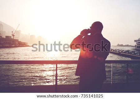 Silhouette of a man leadership dressed in suit is drinking take way coffee, while is standing outside the company against sea sunset background with copy space for your advertising text message