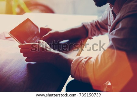 Cropped shot view of hipster guy holding touch pad while sitting at wooden table in modern space interior, young businessman using digital tablet for distance job during free recreation time