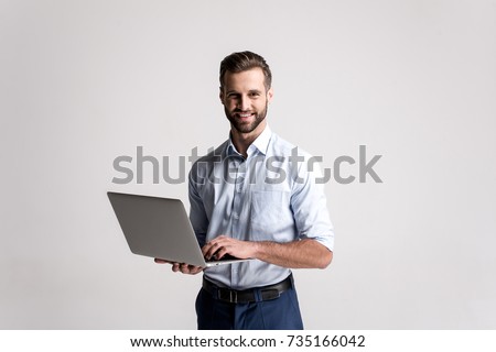 Working with joy. Handsome young man using his laptop and looking at camera with smile while standing against white background.