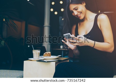 Attractive female student is reading laughable article in network on mobile phone while is sitting in university cafe. Woman chatting with her friends by cell telephone during breakfast in coffe shop