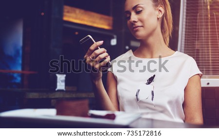 Charming European female browsing wifi via mobile phone during morning breakfast in cafe bar, beautiful woman reading text message on her cell telephone while sitting in coffee shop during free time 