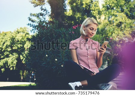 Young female reading pleasant text message from her boyfriend on mobile phone while sitting with book in the park, woman is watching video in internet via cell telephone while she is relaxing outside