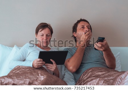 Bored couple, husband and wife in bedroom using personal electronics devices