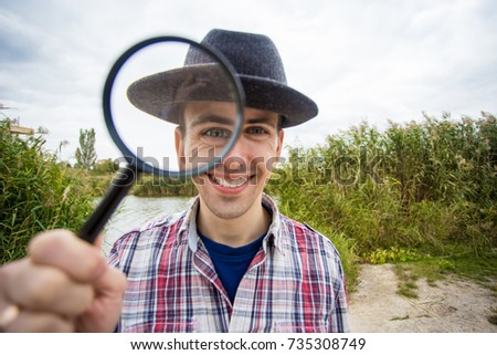A cheerful young man with a funny face in a hat holds a magnifying glass.