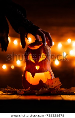 Photo of halloween pumpkin cut in shape of face with witch's hand on background with burning yellow lights