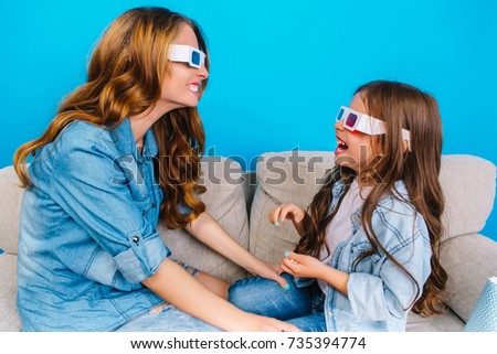Happy mother and her little daughter in jeans clothes smiling to each other on couch isolated on blue background. Wearing 3D glasses, having fun together mum and kids, happiness in childhood