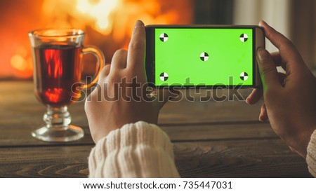 Closeup image of young woman holding mobile phone with green screen next to cup of tea and burning fireplace