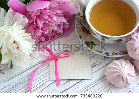 Peonies flowers with card marshmallow pink cup of tea on a white wooden background - stock image.