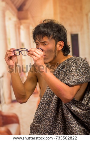 Close up of funny prehistoric man doing funny faces to the camera, holding in his hands a glasses, in a blurred background