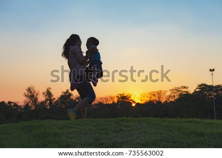 Silhouette of happy little girl child running into the arms of his loving mother for a hug, in front of the sunset in the sky on a summer day on Chatuchak Park at Bangkok City, Thailand.