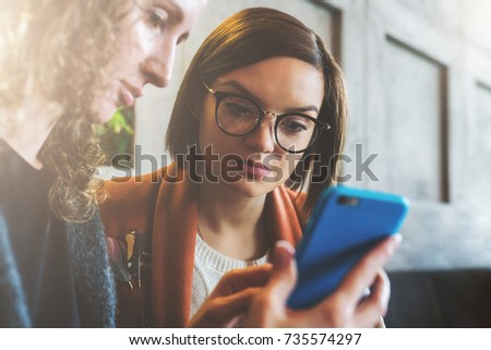Two young women sit in a cafe at the table and use a smartphone. The girl shows her friend a picture on the phone screen. Social media, network. Girls working together, blogging, checking email.
