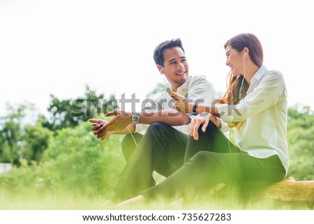 Young Asian lovely couple or college students sit listening to song music on smartphone together in park. Leisure activity, Online love, internet dating app technology, or casual lifestyle concept