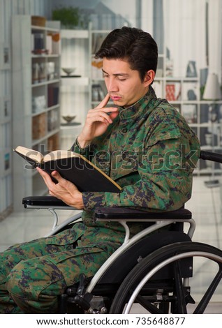 Close up of a young soldier sitting on wheel chair reading a book at home, in a blurred background