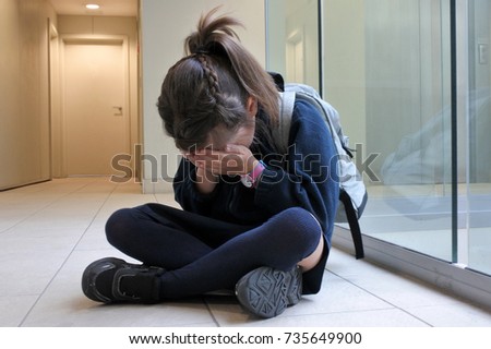 One young elementary school girl wearing school uniform and backpack sitting on a corridor floor crying. Childhood and education concept. Real people. Copy space Royalty-Free Stock Photo #735649900