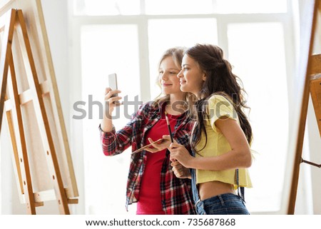 art school, technology and people concept - happy student girls or artists with smartphone, easel, palette and paint brush painting and taking selfie at studio