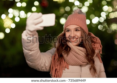 holidays and people concept - beautiful happy young woman taking selfie over christmas tree lights in winter evening