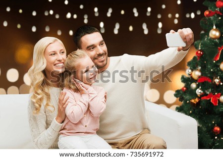 christmas, holidays, technology and people concept - happy family sitting on sofa and taking selfie picture with smartphone over lights background