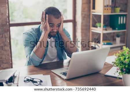 Frustrated young african entrepreneur with sad grimace in front of his laptop in office, noticed a big mistake he did, holding his face with arms palms, wearing jeans casual smart Royalty-Free Stock Photo #735692440