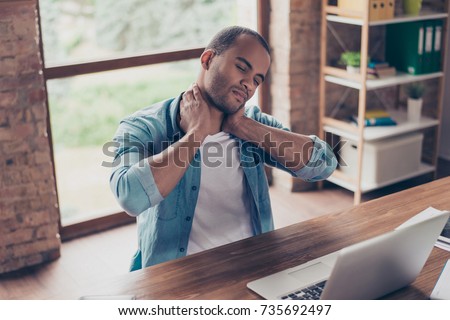 Young frustrated afro american freelancer is having a strong pain in neck. He is at home office, sitting at his modern work station, suffering, massaging it Royalty-Free Stock Photo #735692497
