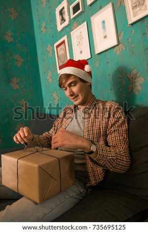 Young adult man wearing red christmas hat wrapping present seated on blue couch in cozy apartment indoor