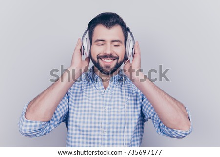 Close up portrait of bearded carefree brunet guy having a break, resting and enjoying the sound, isolated on a light background, touching big modern white ear phones, in blue checkered shirt