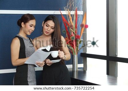 Two Asian Office Girls talk chit chat on paper document or project and drink coffee white cup in blue office room white windows and flower