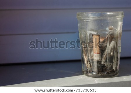 In a dirty glass cigarette butts and ashes. Gray background, side lighting. Harm of smoking, addiction, a fallen man.