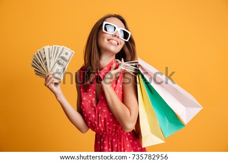 Photo of cheerful elegant young woman in sunglasses and red dress holding fan of money and colorful shopping bags, looking aside, isolated on yellow background