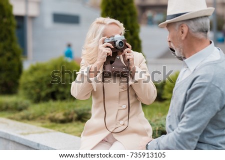 An elderly couple is sitting on the edge of a flower bed. They take pictures of each other on a vintage film camera. They are posing and smiling