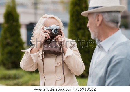 An elderly couple is sitting on the edge of a flower bed. They take pictures of each other on a vintage film camera. They are posing and smiling