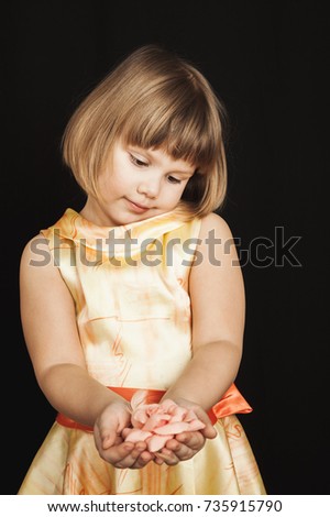 Little Caucasian girl in yellow dress with pink rose flower made of feathers on black background, vertical studio portrait