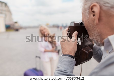 An elderly couple is walking around the square. A woman poses with a dog in her arms, a man takes pictures of her on a film camera. Nearby is a lilac travel bag