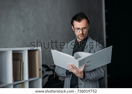 serious businessman in eyeglasses holding folder and working with papers in office