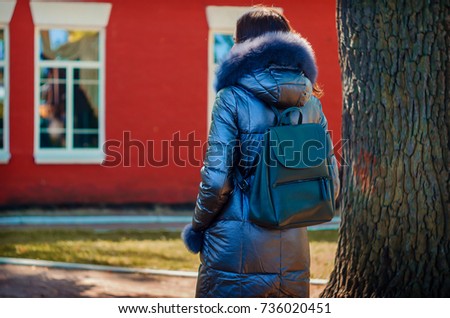 beautiful slender brunette woman stands under a tree in a park next to a red house in a winter jacket (down jacket) with a gray city backpack on her shoulders